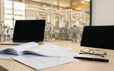 Engineering desk with datasheets overlooking a production hall (AI-generated illustration)